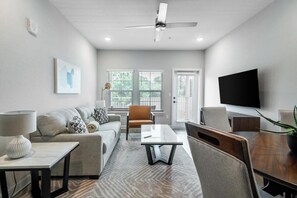 Ein modernes Wohnzimmer mit einer Couch, einem Couchtisch und Sesseln, einem an der Wand befestigten Flachbildfernseher sowie einem Deckenventilator.