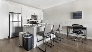 Een moderne keuken met witte kasten, roestvrijstalen apparatuur, een klein eiland met zitplaatsen, en een bureau met een stoel.