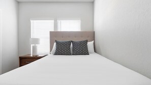 Quarto com cama, duas almofadas estampadas, abajur e mesa de cabeceira.