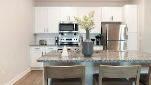 A modern kitchen with white cabinets, a stainless steel refrigerator, a microwave, and a stove. There is a granite countertop and two wooden chairs.