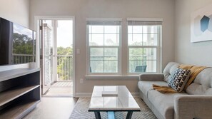 A living room with a TV, a sofa, a coffee table, and two windows. - Landing | Amazing 1BD, Gym, Pool (Palm Bay)