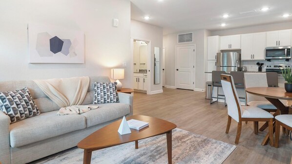 A modern living room with a sofa, coffee table, and dining area with chairs and a table. - Landing | Spectacular 2BD, Pool, Gym (Palm Bay)