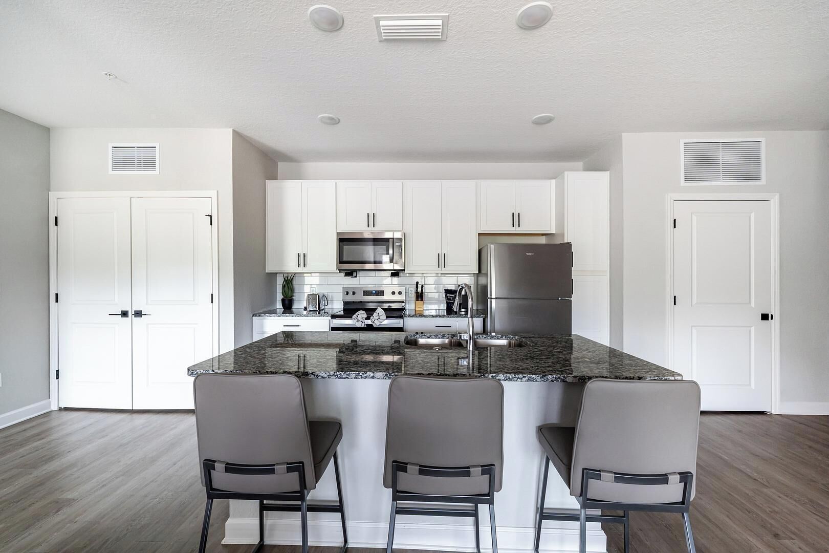 A modern kitchen with white cabinets, a stainless steel refrigerator, a microwave, and a granite countertop. There are three bar stools and a potted plant.
