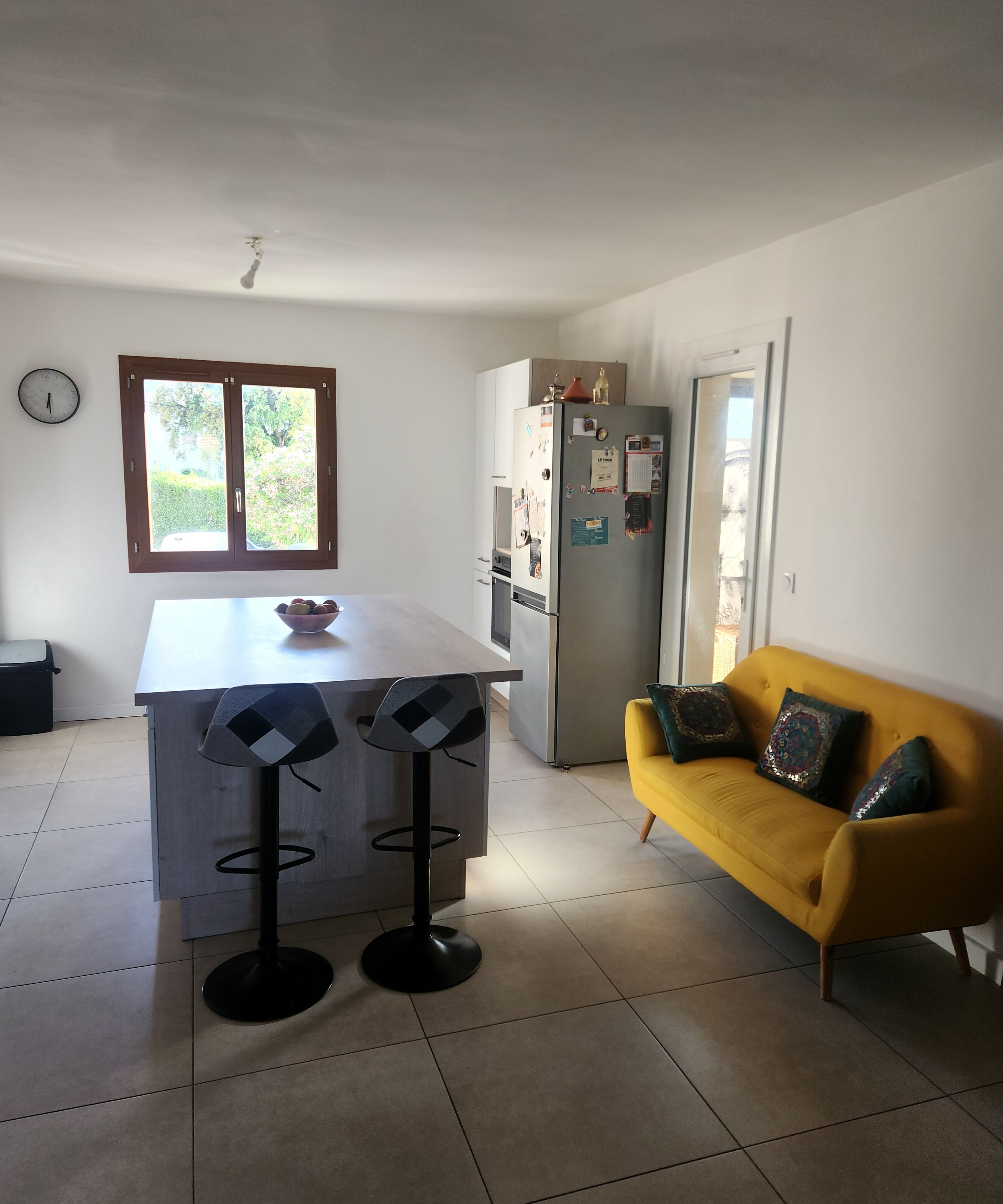 A kitchen with a central island, a yellow sofa, and a window with a view of greenery.
