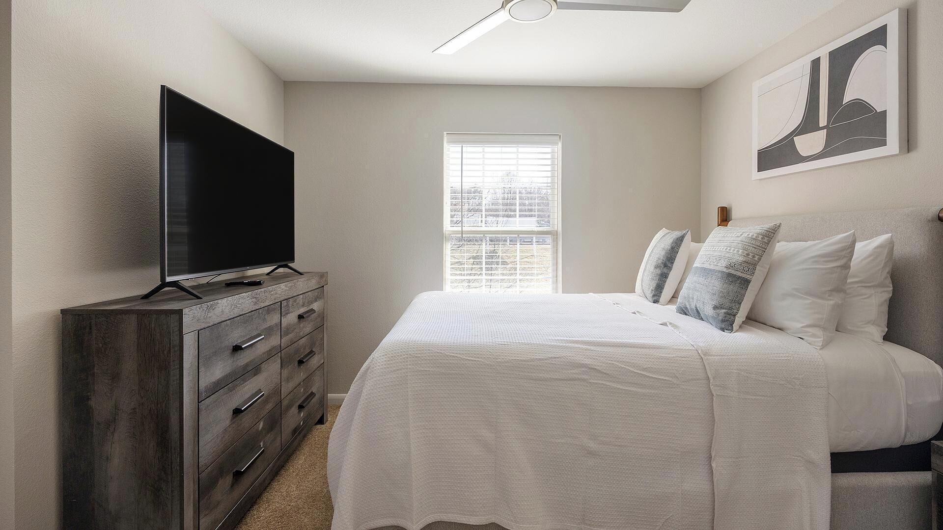 A bedroom with a bed, a dresser, a television on top of the dresser, a window, and a ceiling fan.