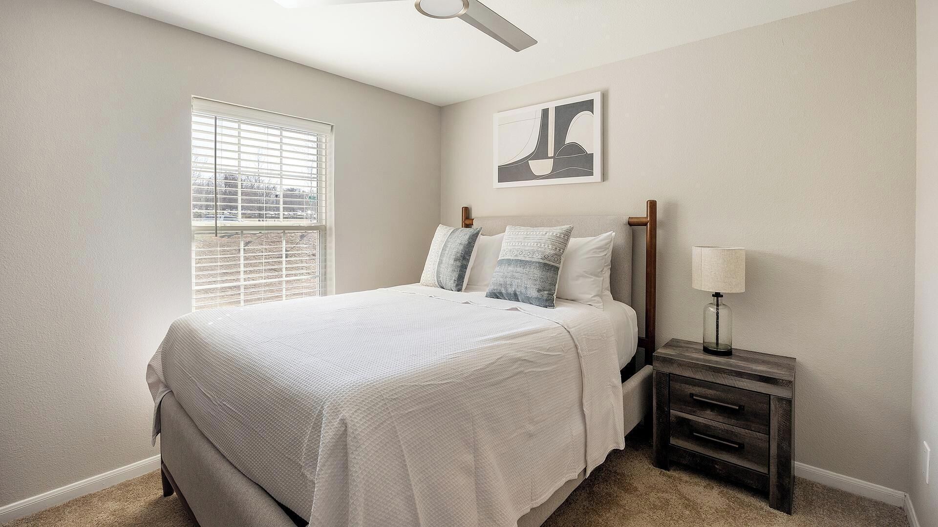 A bedroom with a bed, a nightstand, a lamp, a window with blinds, and a ceiling fan.