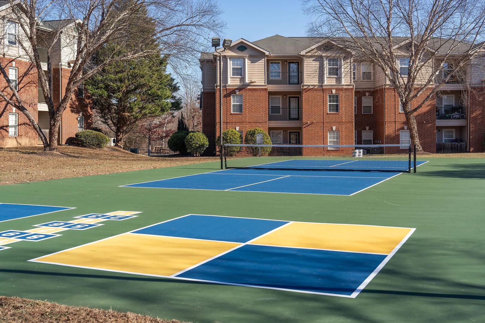 A tennis court with a net, surrounded by brick buildings and trees.