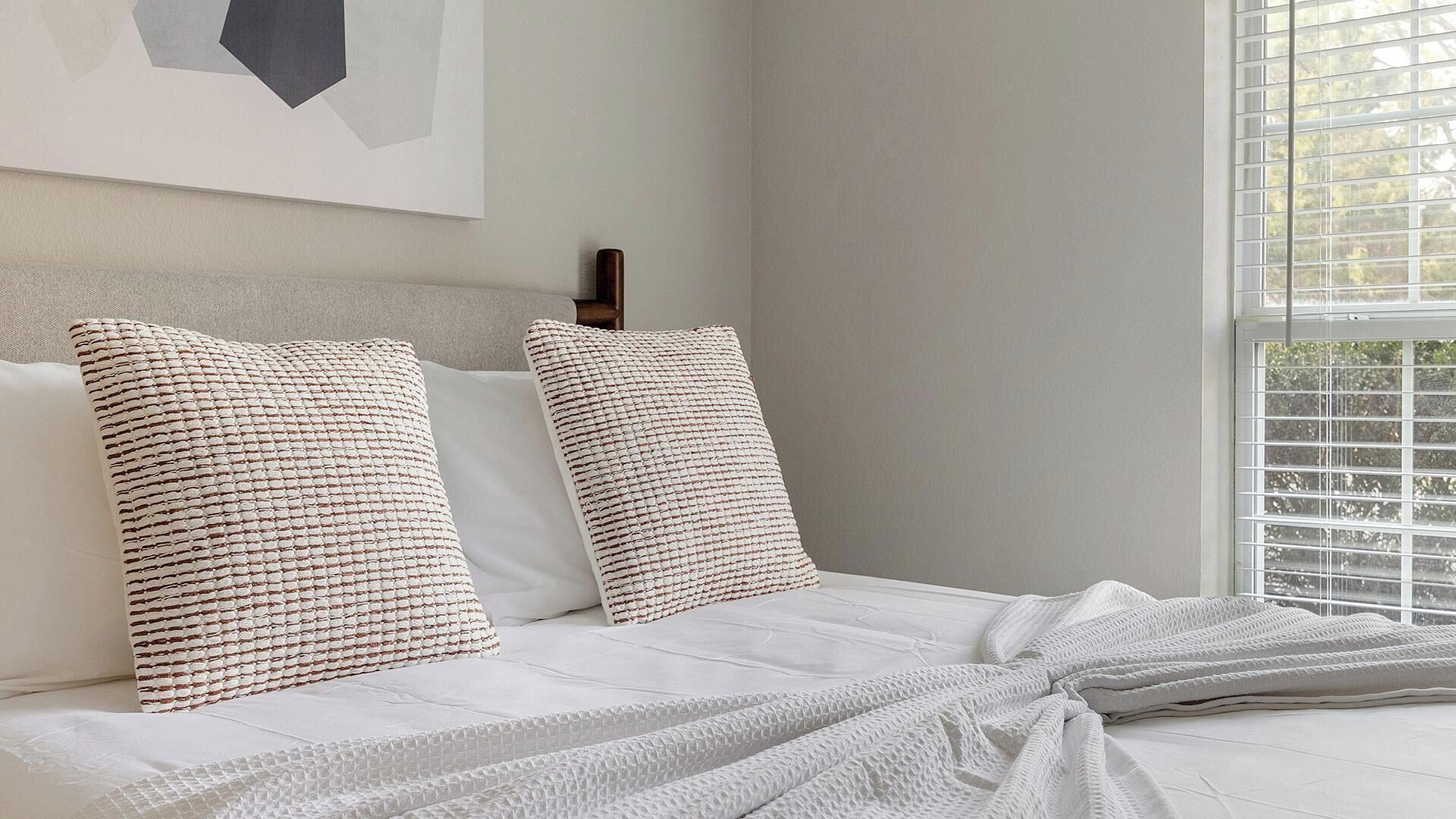 Un lit avec du linge de lit blanc, deux oreillers à texture et une fenêtre avec des stores.