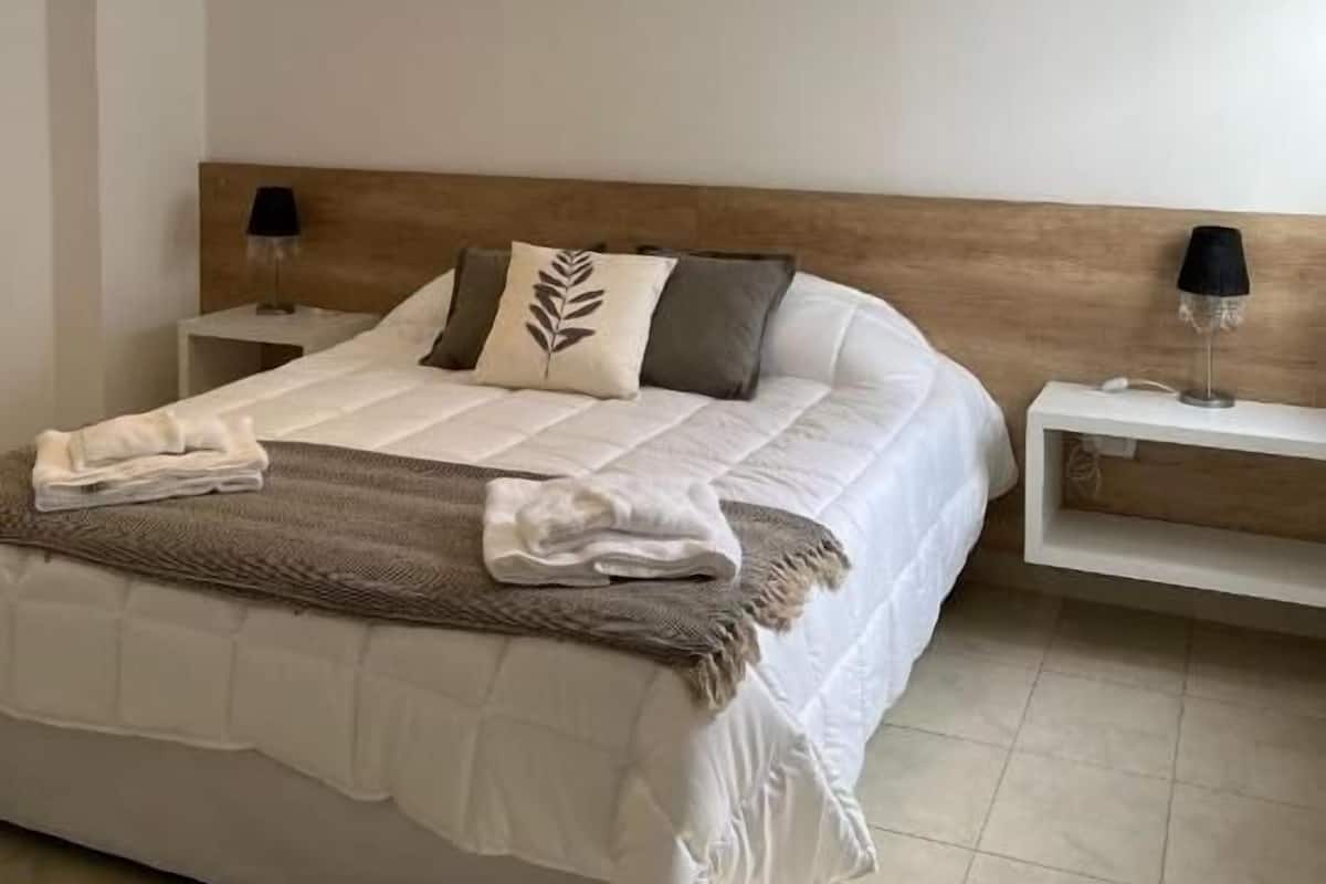 A modern bedroom with a wooden headboard, a neatly made bed, a bedside table with a lamp, and a small shelf.