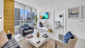 A modern living room with a large window, a flat-screen TV, and a view of a cityscape. - LUXORA 2BR 1 BA 16TH FLOOR W -FREE SPA (Miami)