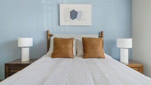 A neatly made bed with white linens, two brown pillows, and a wooden headboard. There are two bedside tables with lamps, and a framed abstract artwork hangs above the bed. - Landing | Modern 2BD in Eastpoint (Baytown)