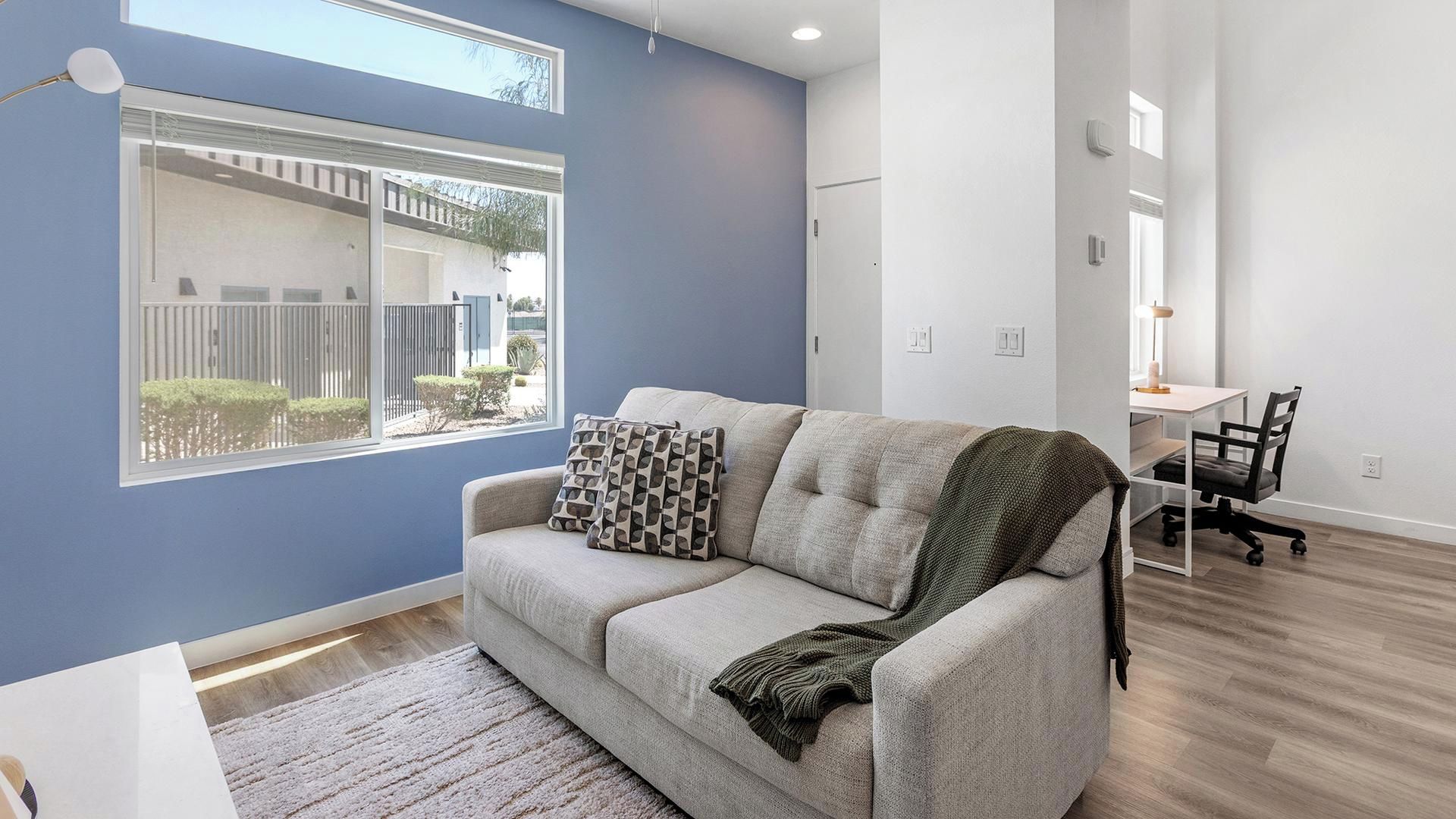 A modern living room with a sofa, a dining table with chairs, and a window with a view of a fence and greenery.