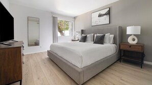 Un dormitorio moderno con una cama grande, una mesita de noche con lámpara, un aparador y una ventana con vista a árboles.