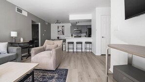 Una sala moderna con un sofá, una mesa de centro y un televisor. Hay una zona de cocina con un microondas y una cafetera.