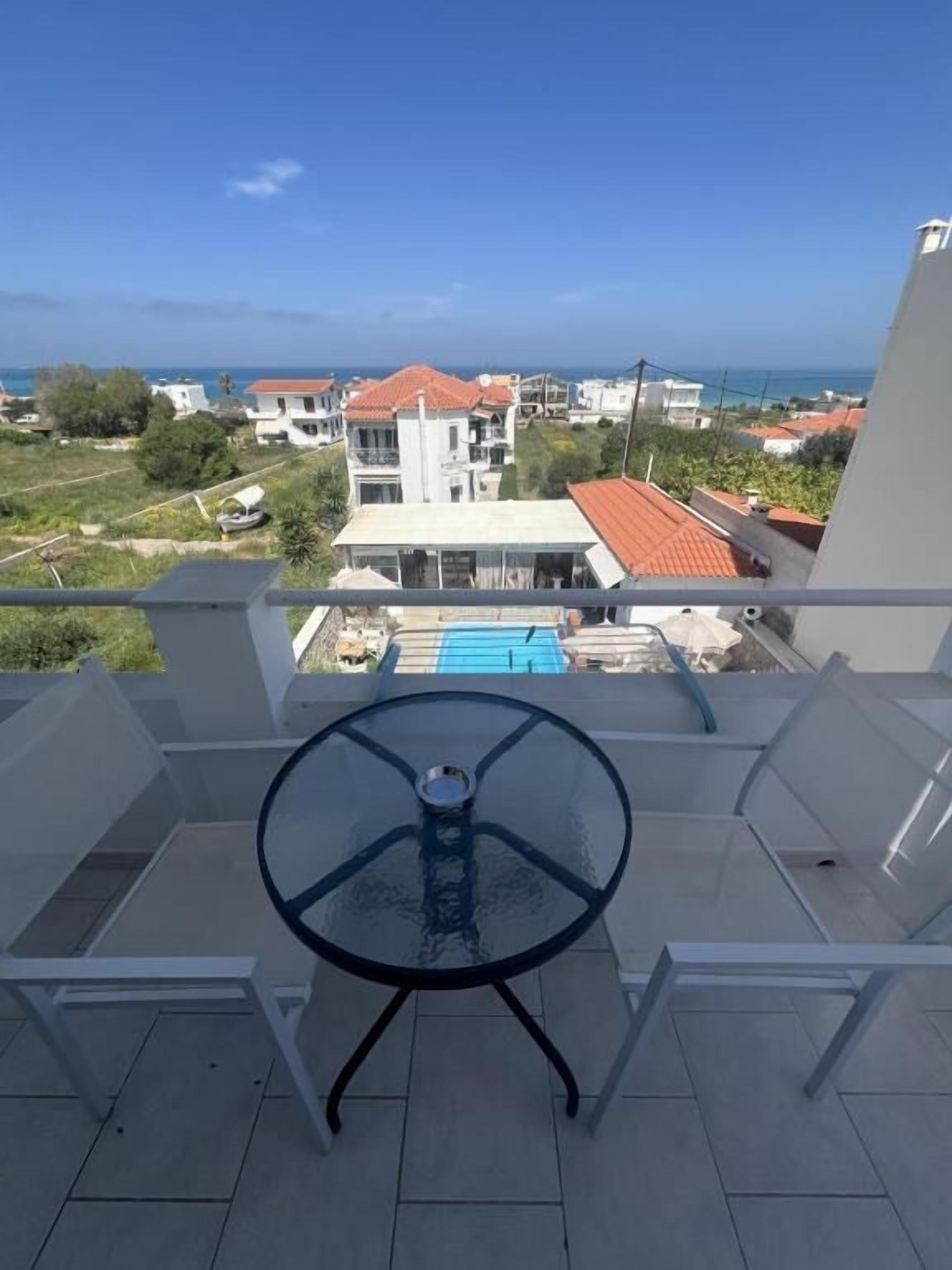 Uma varanda com mesa e cadeiras de vidro, com vista para uma área residencial com casas e uma piscina azul.