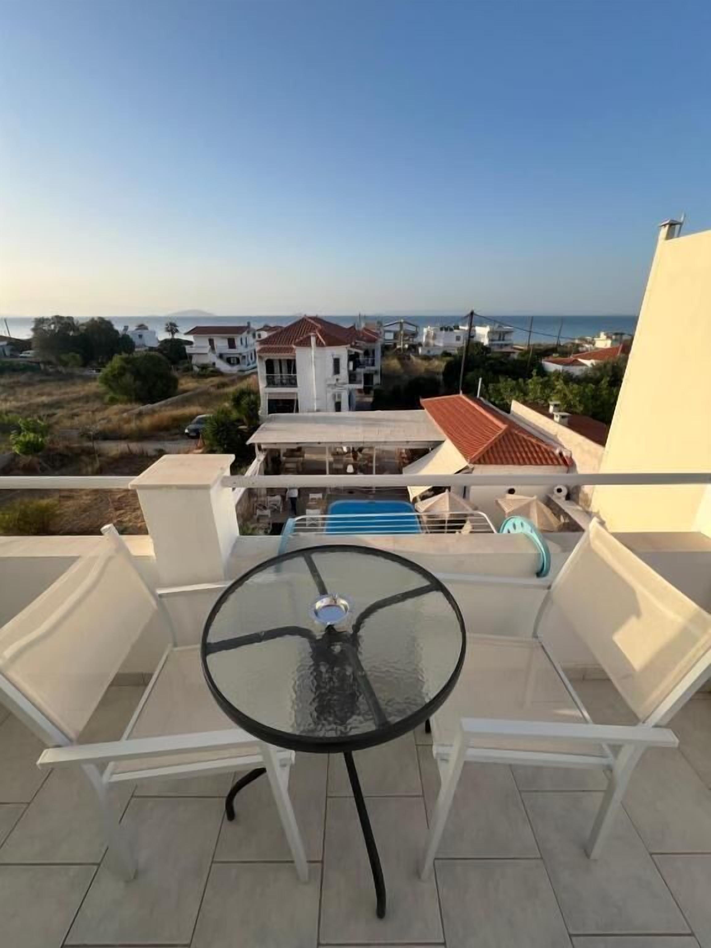 A balcony with a glass table and chairs, overlooking a residential area with houses and a clear sky.