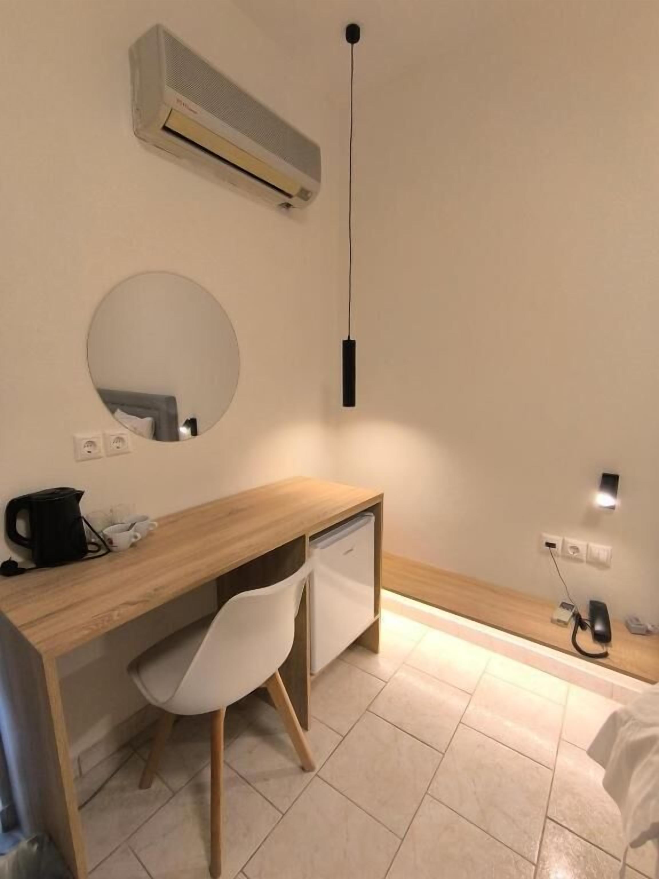Ein Hotelzimmer mit einem Holztisch, einem weißen Stuhl, einem runden Spiegel, einer Klimaanlage und einem Bett mit weißer Bettwäsche.