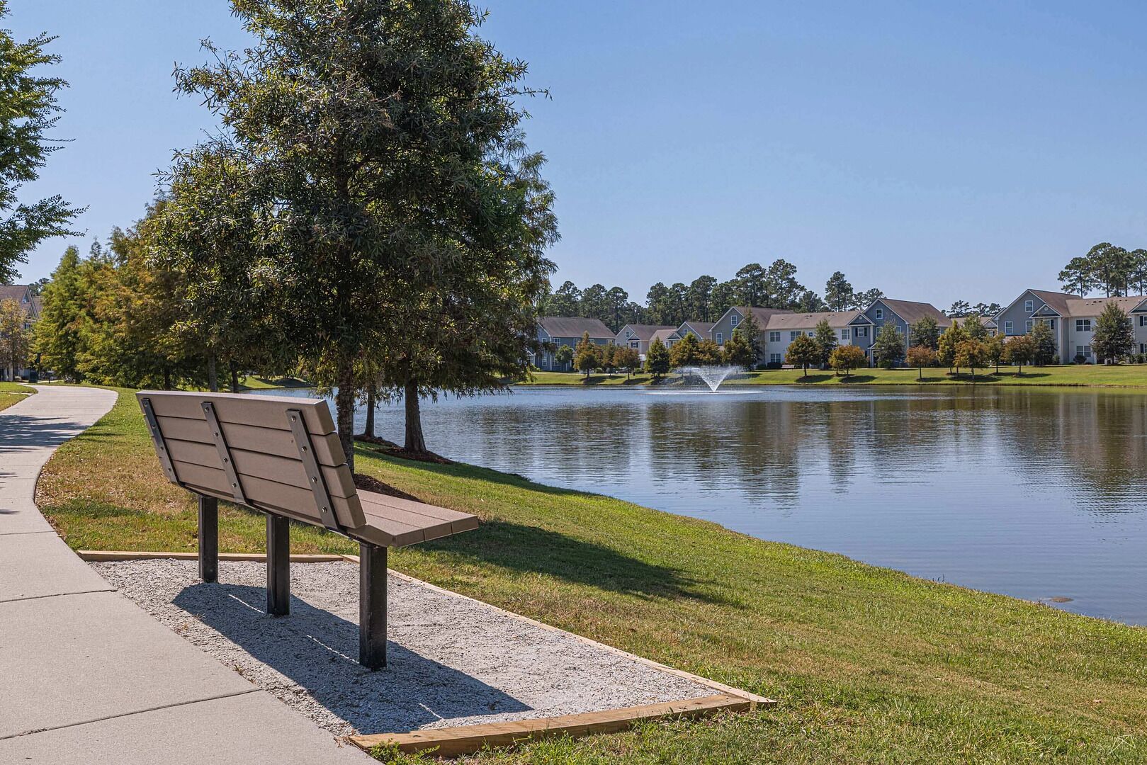 A bench by a lake with a fountain, surrounded by grass and trees, and residential buildings in the background.