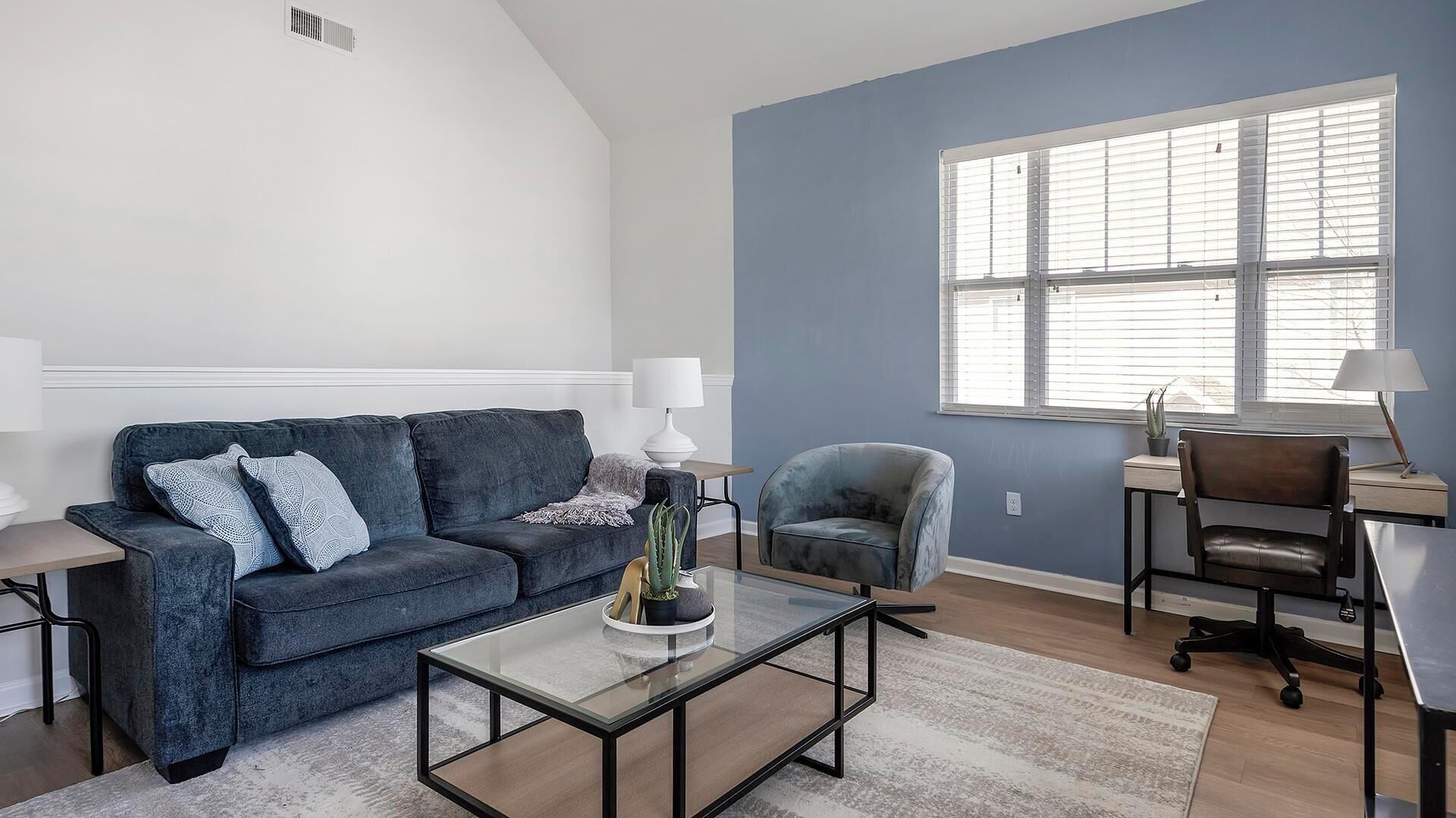 Un salon avec un canapé bleu, une table basse en verre, un fauteuil bleu et un bureau avec une chaise.