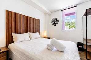 A neatly made bed with a wooden headboard, a bedside table, a wall-mounted picture frame, and a window with a view. - Semillero Obispado (Monterrey)