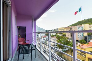 A balcony with purple walls, a railing, and a view of a cityscape with buildings and a flag. - Semillero Obispado (Monterrey)