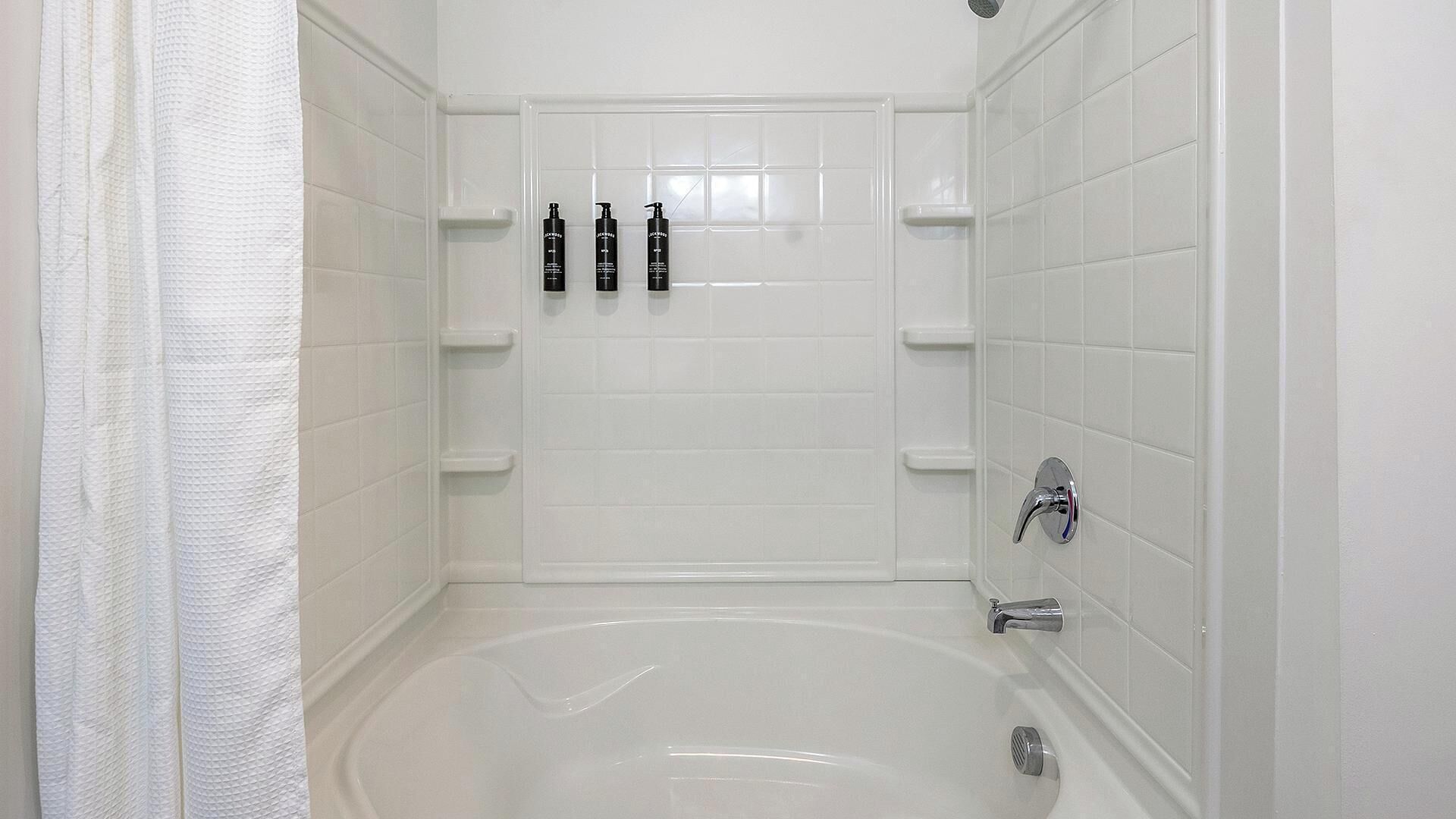 A modern bathroom with a bathtub, white tiled walls, and three black bottles on a shelf.