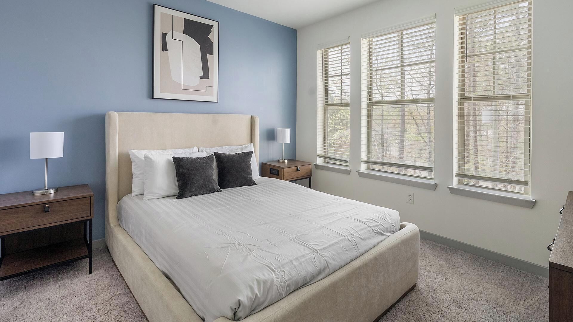 Une chambre à coucher comprenant un lit, deux tables de chevet, une lampe, un tableau encadré et trois fenêtres équipées de stores.