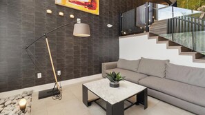 Una sala moderna con sofá, una mesa de centro, una lámpara de pie y una escalera con barandillas de vidrio.