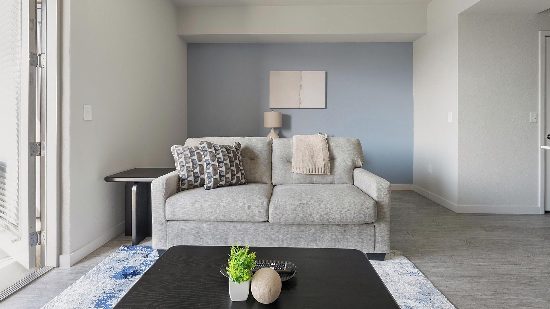 A living room with a grey sofa, a small side table, a black coffee table with decorative items, and a blue and white patterned rug.