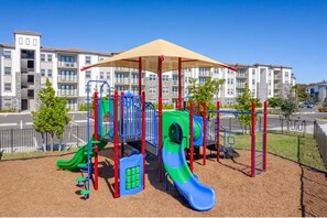 A playground with slides and climbing structures, surrounded by a fence and a building in the background. - Landing | Modern 2BD, Clubhouse, Gym (Fort Myers)