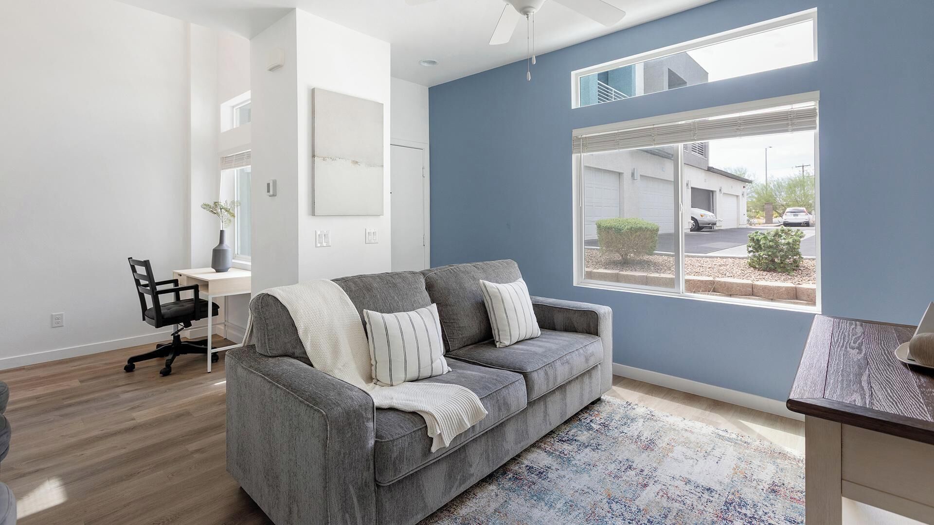 A modern living room with a grey sofa, a desk, and a large window with a view of a residential area.