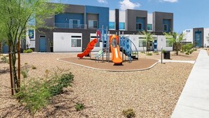 Ein Spielplatz mit Rutschen und Klettergerüsten, umgeben von einem Kiesbereich und einem Gebäude mit mehreren Fenstern und Türen.