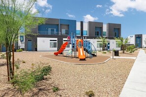 A playground with slides and climbing structures, surrounded by a gravel area and a building with multiple windows and doors. - Landing | Stunning 2BD, Gym (North Las Vegas)