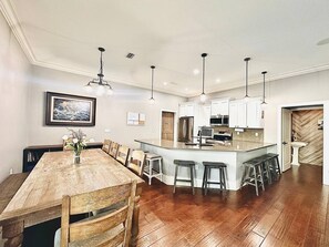 A spacious kitchen with white cabinets, a central island with stools, and a dining area with a wooden table and chairs. - Luxury Beach Retreat - Sleeps 14 - 5BR - Community Pool - Minutes to Beach (Miramar Beach)