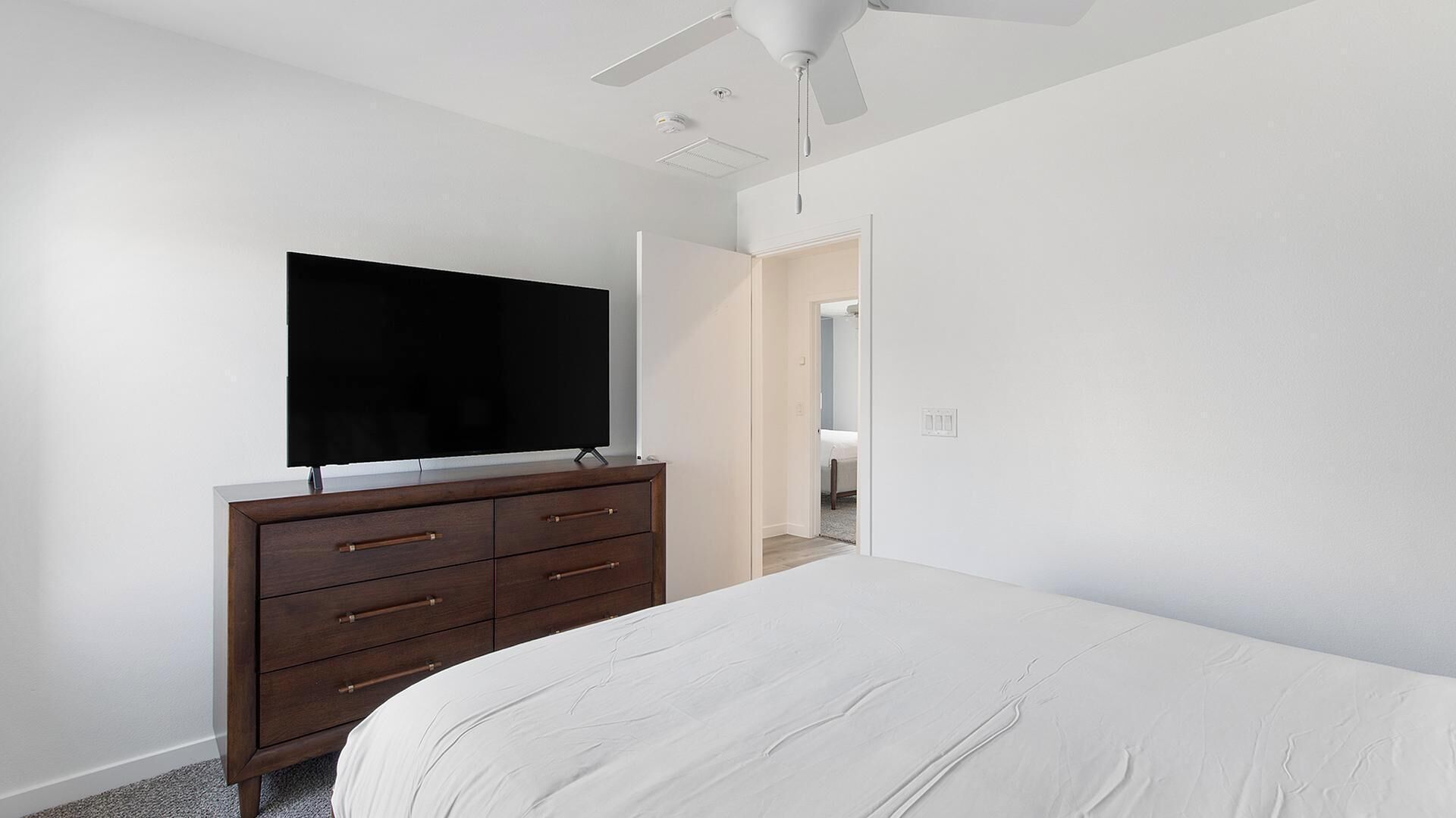 A bedroom with a bed, a dresser, a television on the wall, and a ceiling fan.