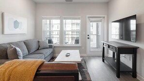 A modern living room with a grey sofa, a coffee table, a TV, and a large window. - Landing | Stylish 1BD, Gym, Pool (Palm Bay)