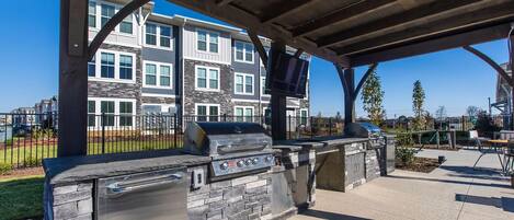 An outdoor barbecue area with a stone grill and a TV mounted on a wooden pergola.