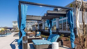 A covered outdoor seating area with blue canopies, a pool, and a building with large windows in the background. - Landing | Beautiful 1BD, Clubhouse, Yoga Studio (Murfreesboro)