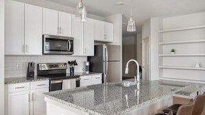 Una cocina moderna con gabinetes blancos, electrodomésticos de acero inoxidable, encimeras de granito e iluminación colgante.