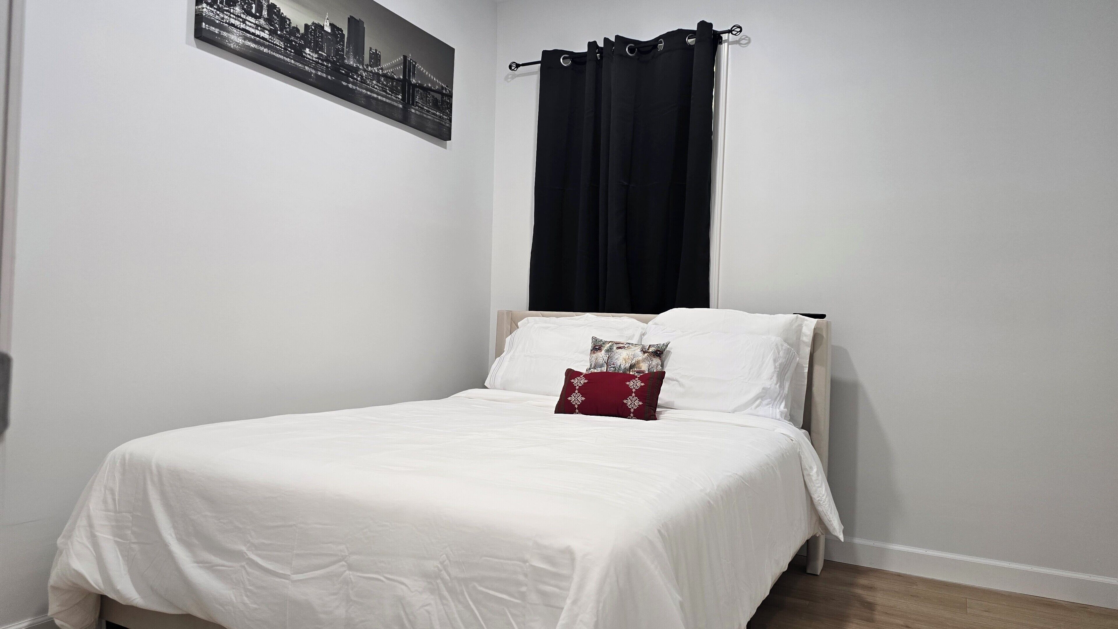 A neatly made bed with white bedding, a black curtain, and a cityscape painting on the wall.