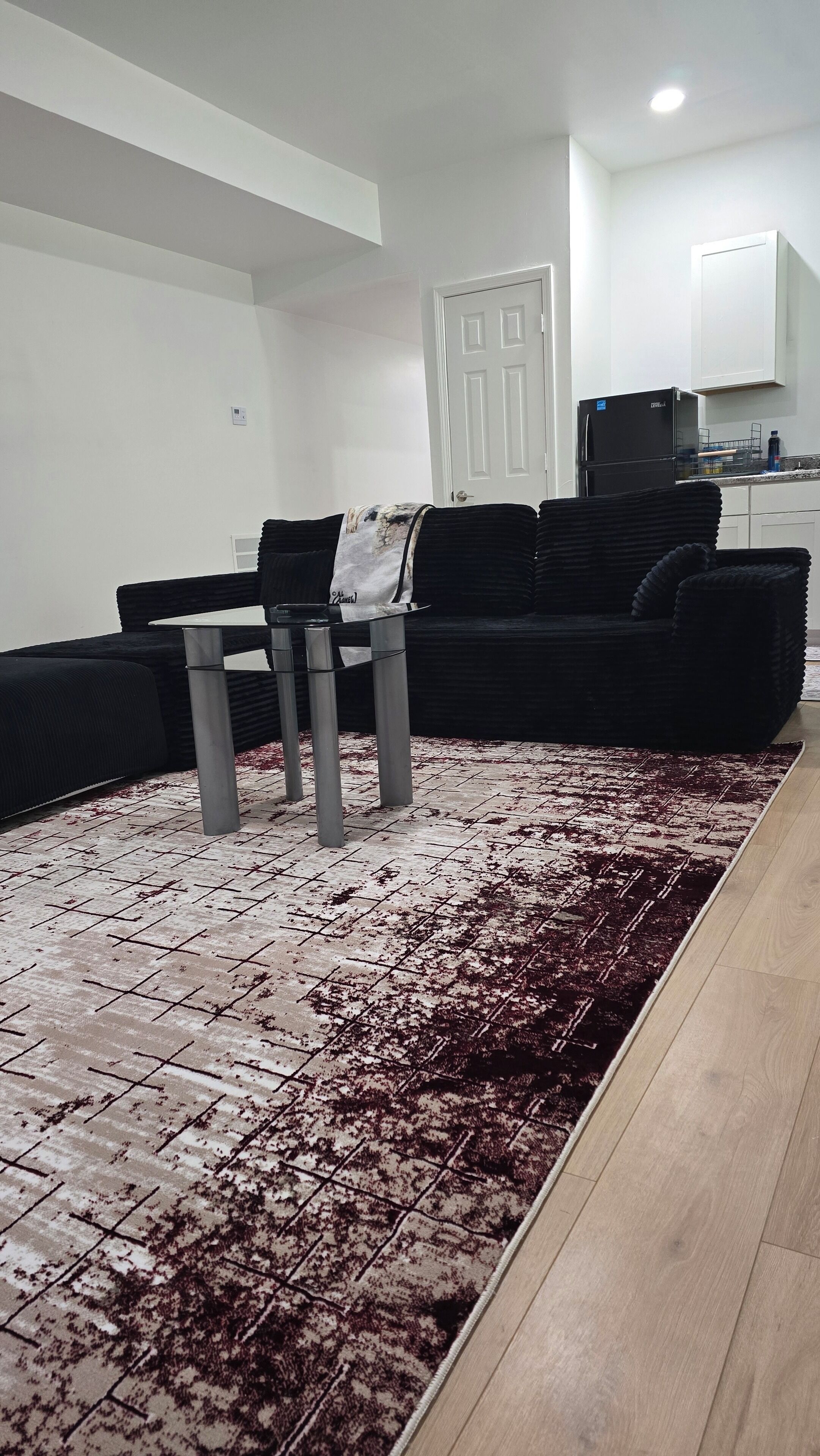 A living room with a black sofa, a glass coffee table, a patterned rug, and a kitchen area in the background.