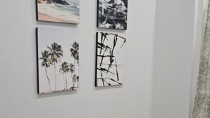A room with a white wall, a wooden floor, and three artworks: a beach scene, a black and white abstract, and a palm tree painting. - 2-bedroom building in charming Stockbridge with cool AC (Stockbridge)