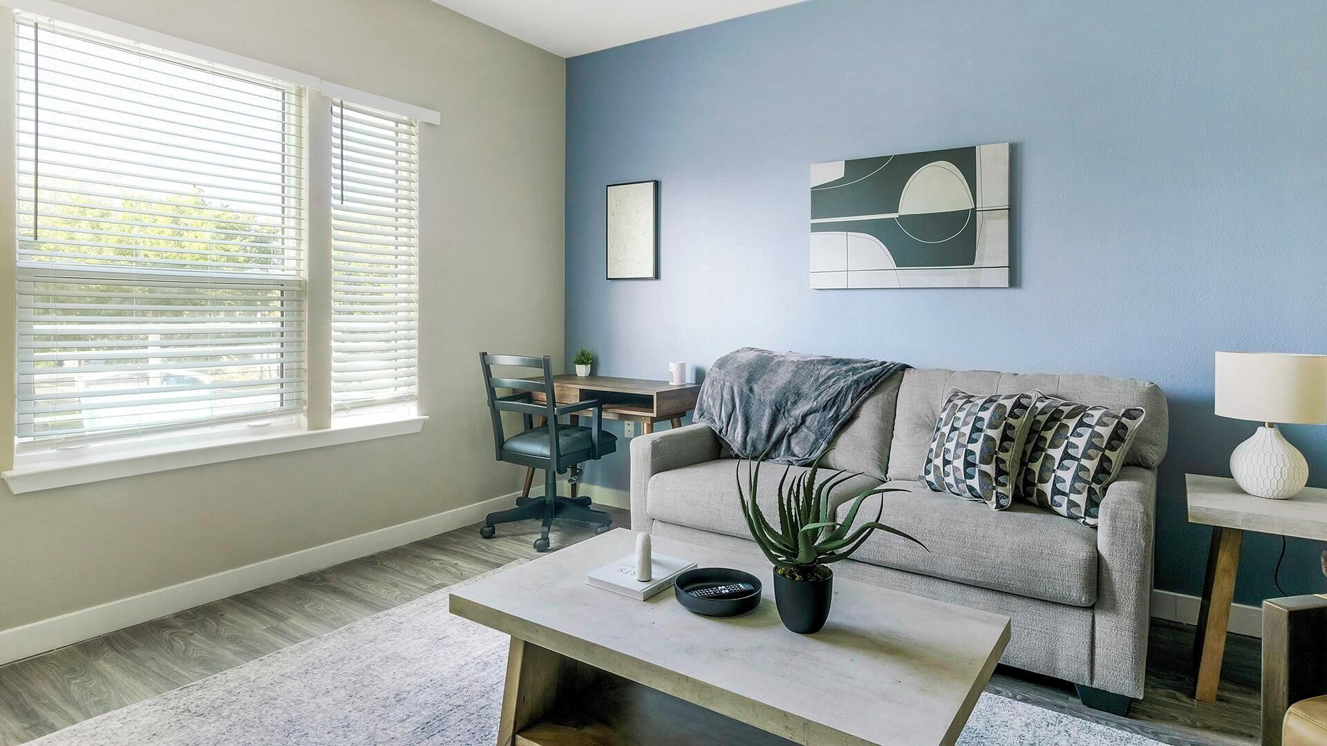 A living room with a sofa, coffee table, and a desk with a chair. There is a window with blinds, a plant on the coffee table, and artwork on the wall.