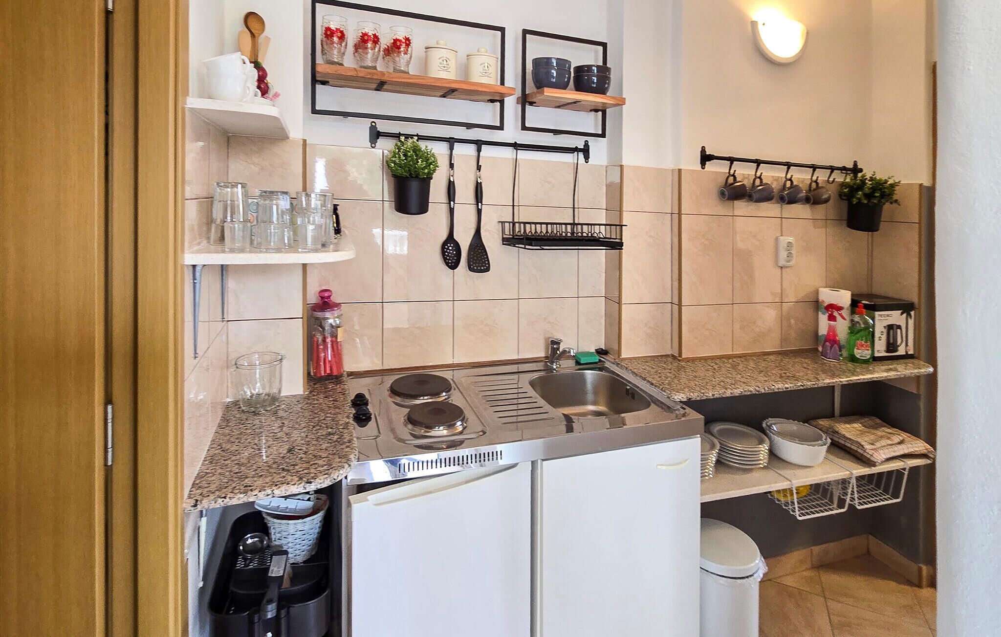 Een keuken met een spoelbak, fornuis en kastjes; planken met glazen en keukengerei; en een wandlamp.