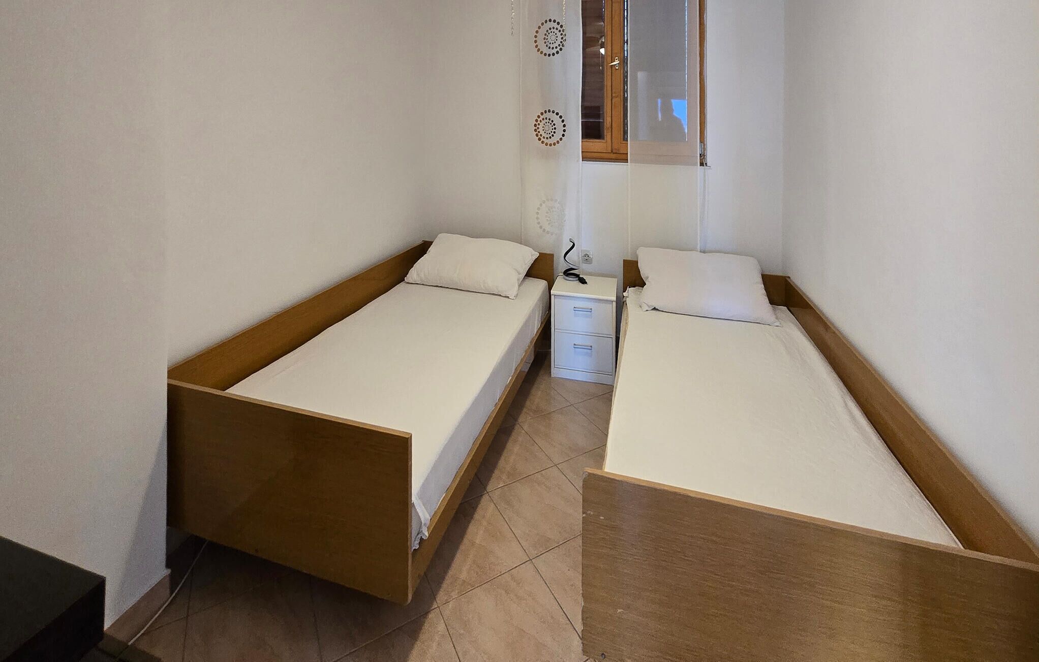 A small, compact room with two single beds, a bedside table with a lamp, and a window with a wooden frame.