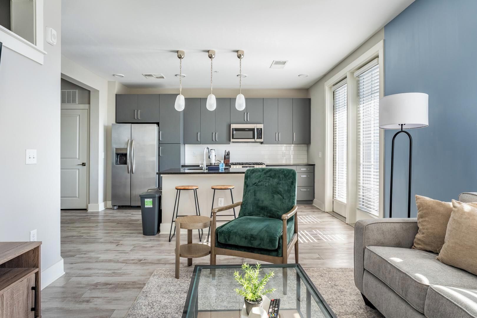 Een moderne woonkamer met een groene fauteuil, een grijze bank, een glazen salontafel en een keuken op de achtergrond.