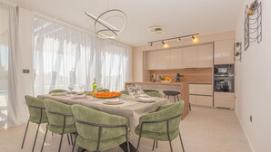 A dining area with a table set for six, green upholstered chairs, a kitchen in the background, and a modern pendant light fixture. - Nice home in Bristani with sauna (Bristani)
