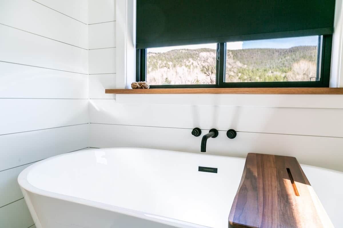 A modern bathroom with a freestanding bathtub, a window with a view of a natural landscape, and a wooden shelf above the tub.
