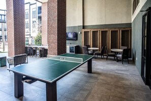 An indoor ping pong table surrounded by chairs and tables, with brick pillars and a TV mounted on the wall. - Landing | Stunning 2BD, Gym, Clubhouse (Lewisville)