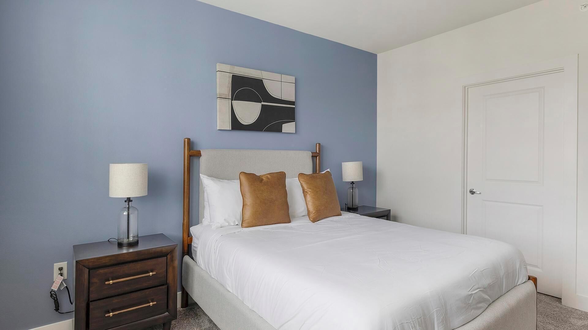 A bedroom with a bed, two bedside tables, a nightstand, a door, and a wall-mounted artwork.